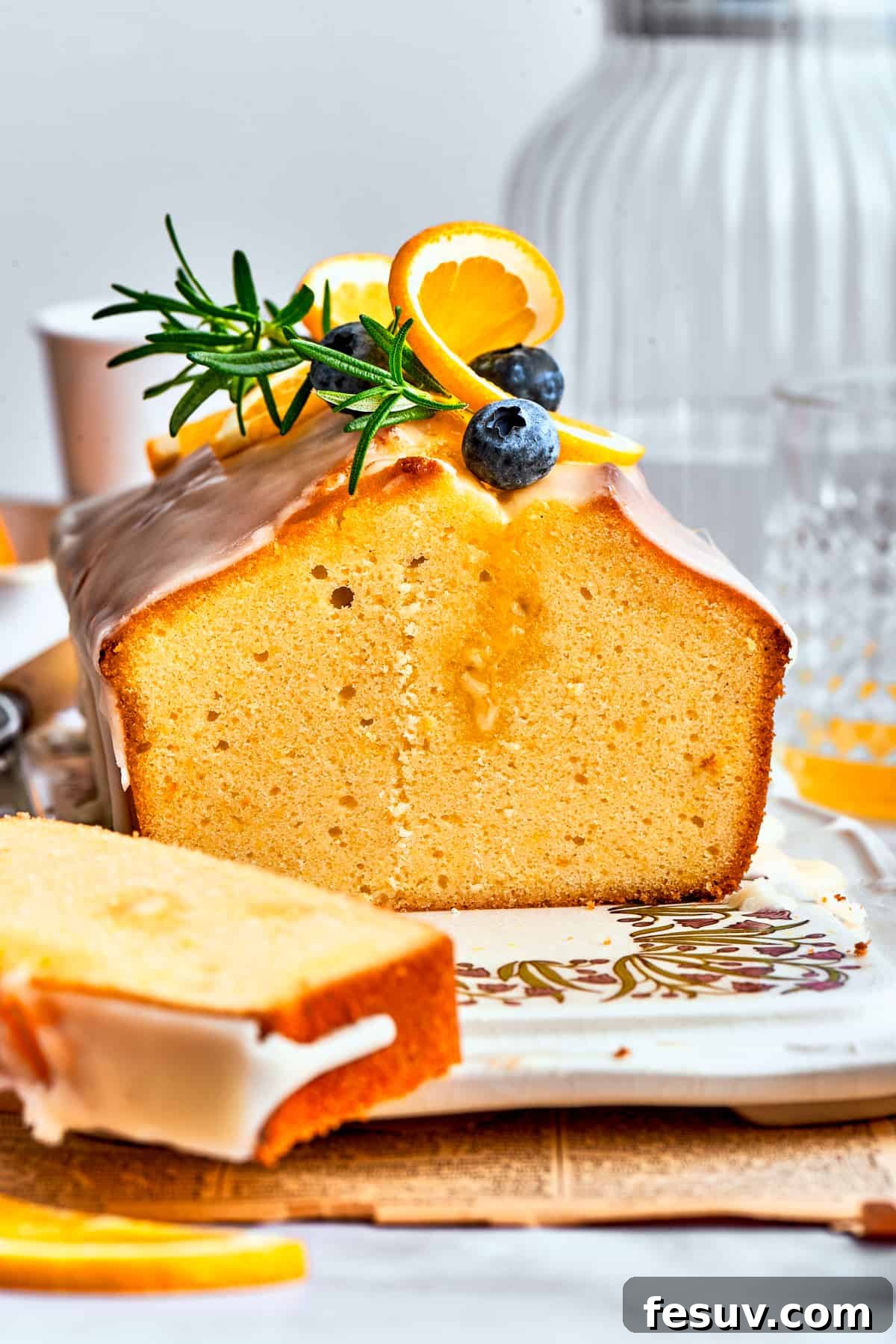 A side view of a slice of orange cake and the entire loaf, topped with blueberries, fresh rosemary, and orange slices. The cake features a gorgeous orange glaze.