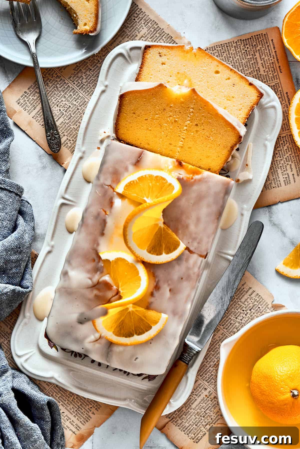 A sliced orange loaf cake topped with orange glaze, orange slices, and wish more oranges surrounding it.