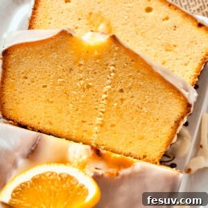 Two slices of glazed orange cake rest atop each other, with the rest of the loaf visible nearby topped with orange slices.
