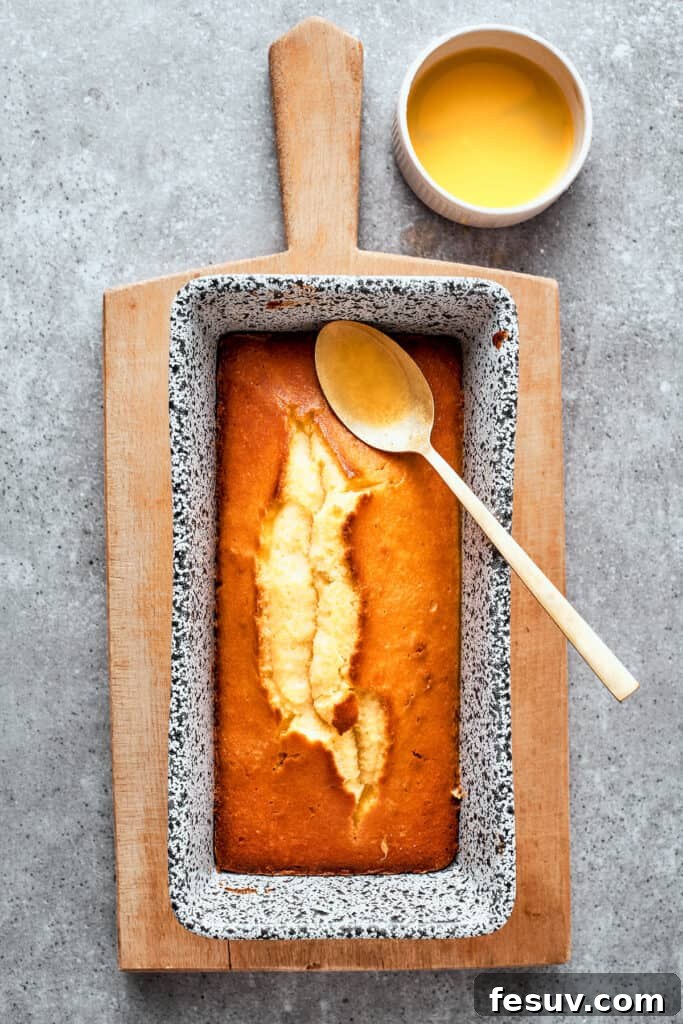 The baked loaf pan of orange cake is shown with a spoon resting on it and a bowl of orange glaze next to it ready to be added to the top.