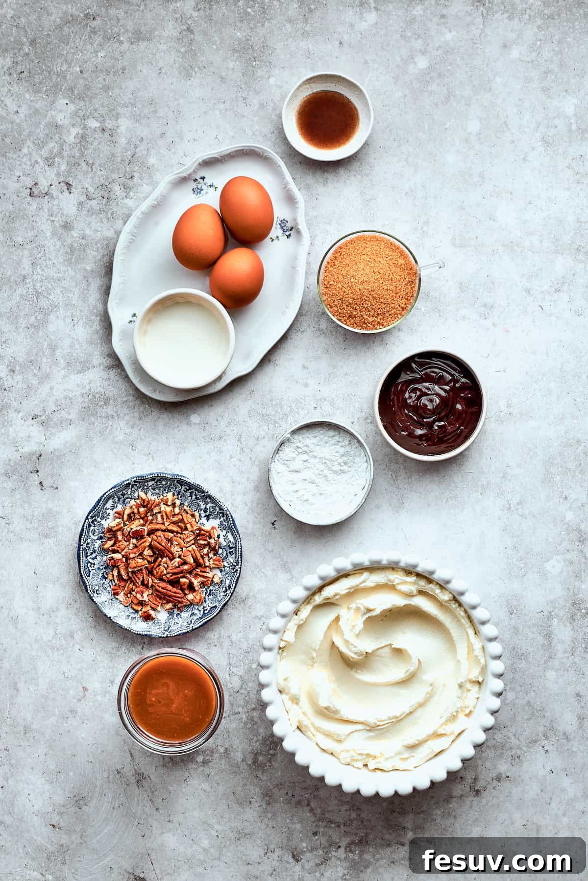 Assorted ingredients for Turtle Cheesecake laid out on a white surface: eggs, pecans, caramel sauce, cream cheese, chocolate chips, brown sugar, vanilla, and sour cream.