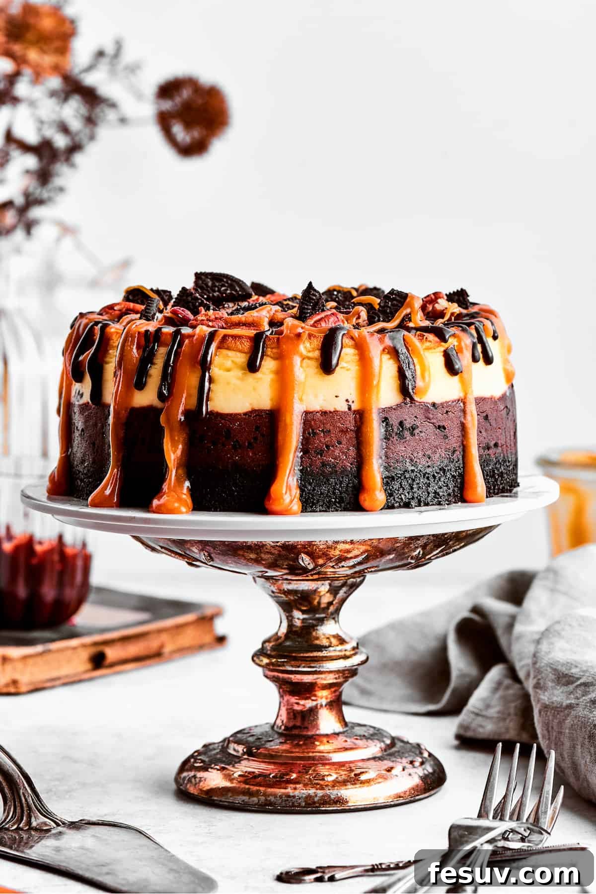 A beautifully crafted Turtle Cheesecake, generously drizzled with caramel sauce, resting on an elegant bronze cake stand.