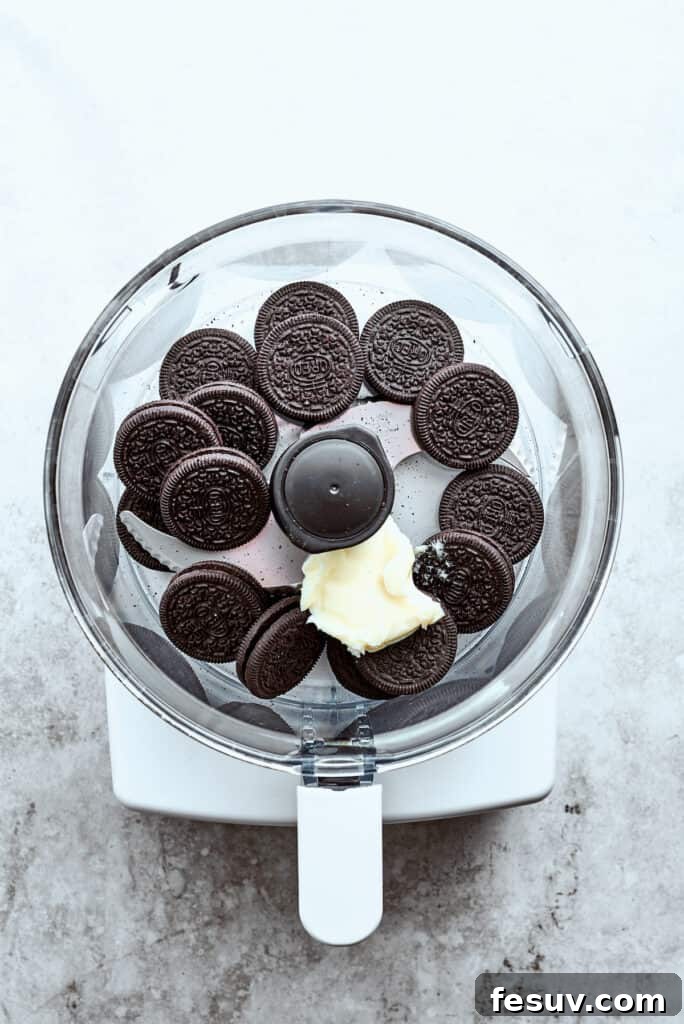 Chocolate sandwich cookies being placed into a food processor alongside a pat of butter, ready to be processed for the cheesecake crust.