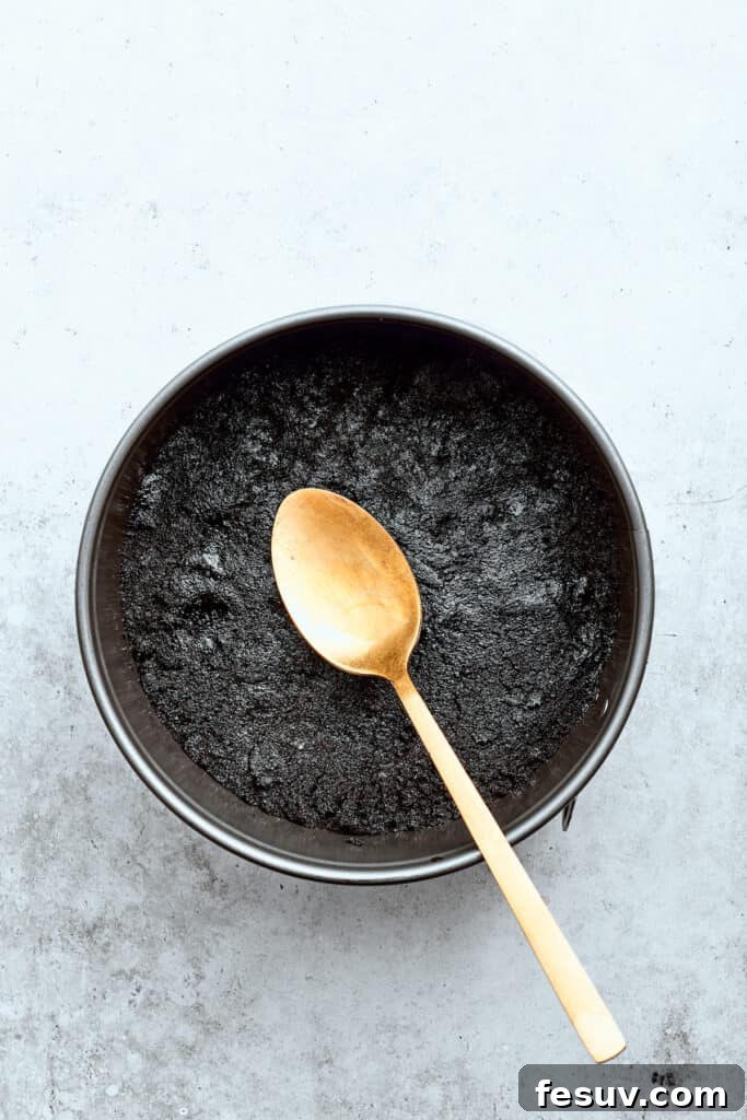 A spoon gently presses a compacted chocolate cookie crust into a 9-inch springform baking pan, ensuring an even and firm base for the cheesecake.