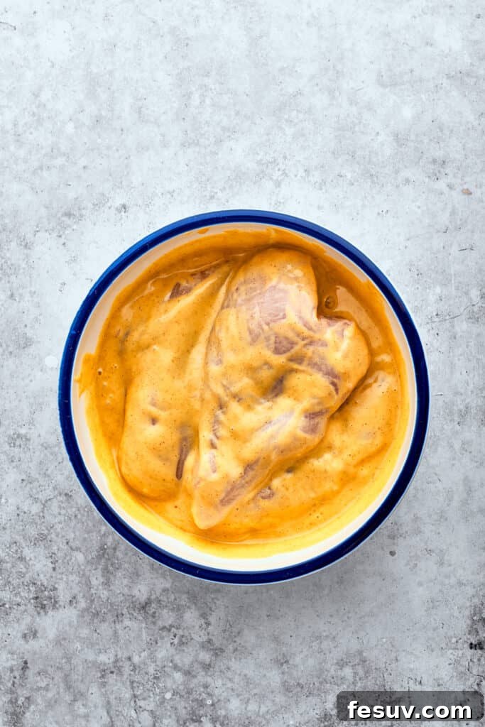 Chicken breasts marinate in a white bowl of yellow honey mustard sauce.