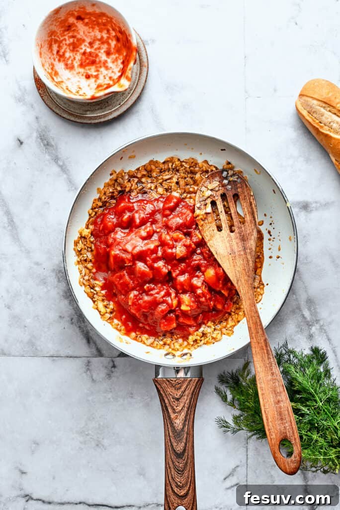 Mixing Tomatoes into the Aromatic Base Thick, rich diced tomatoes are thoroughly stirred into a white skillet, combining with the fragrant base to form the hearty sauce.