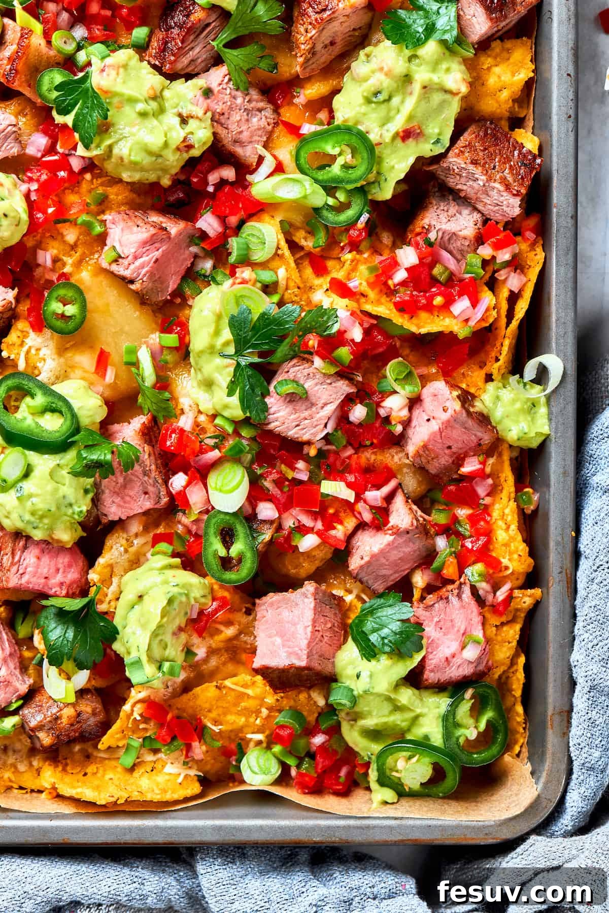 A corner of a pan of steak nachos is shown colorfully topped with guacamole and tomatoes.