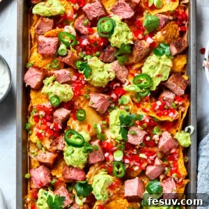A baked pan of steak nachos is topped with dollops of guacamole and chunks of steak.