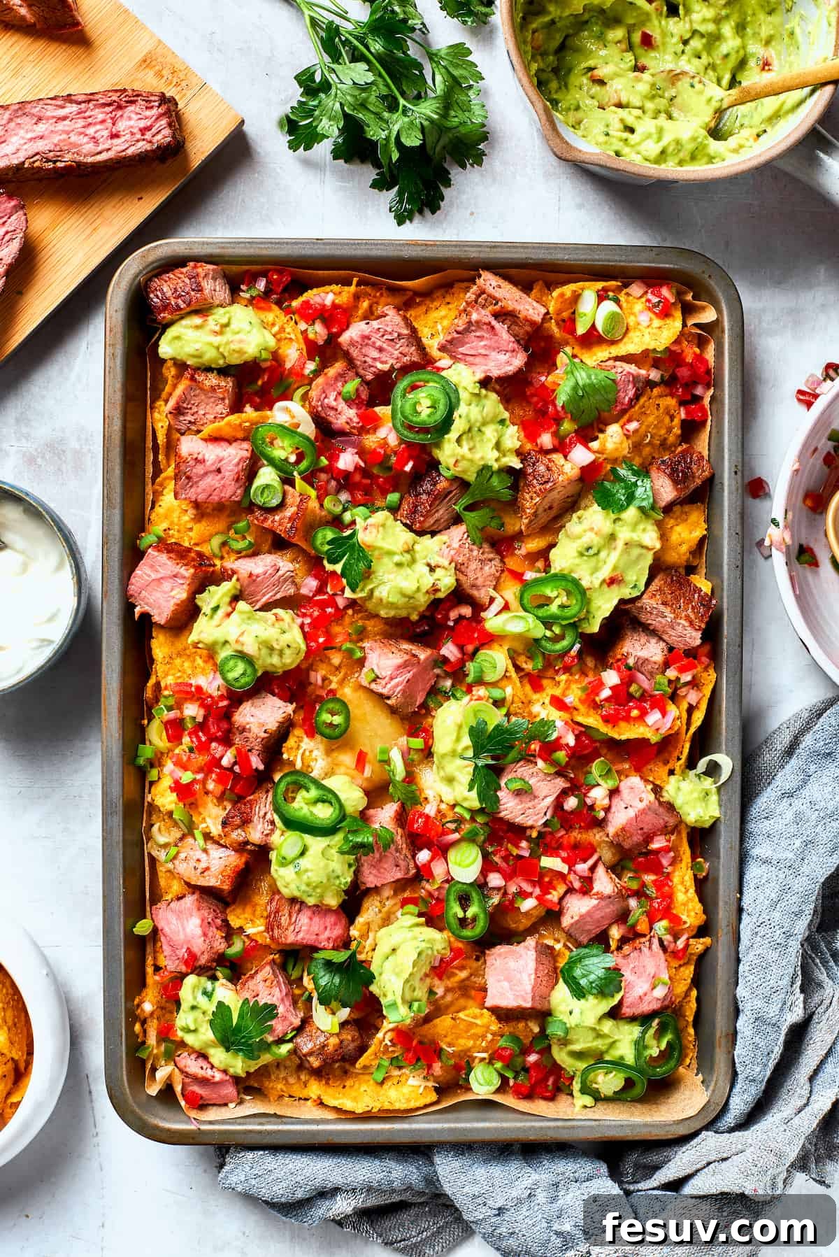 A metal baking sheet of steak nachos is surrounded by ore steak and toppings.