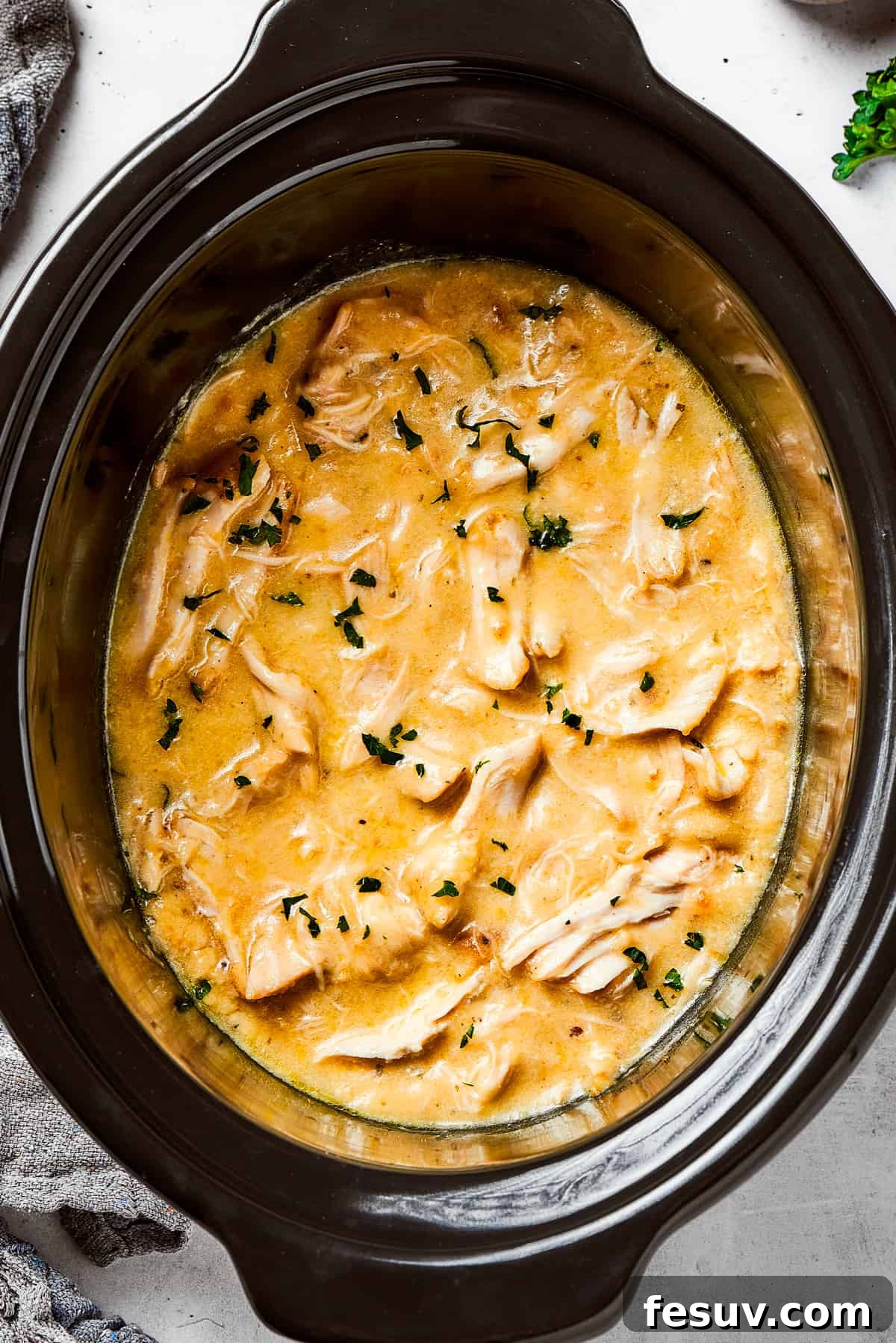 Overhead image of Shredded chicken in a creamy sauce cooking in a black slow cooker.