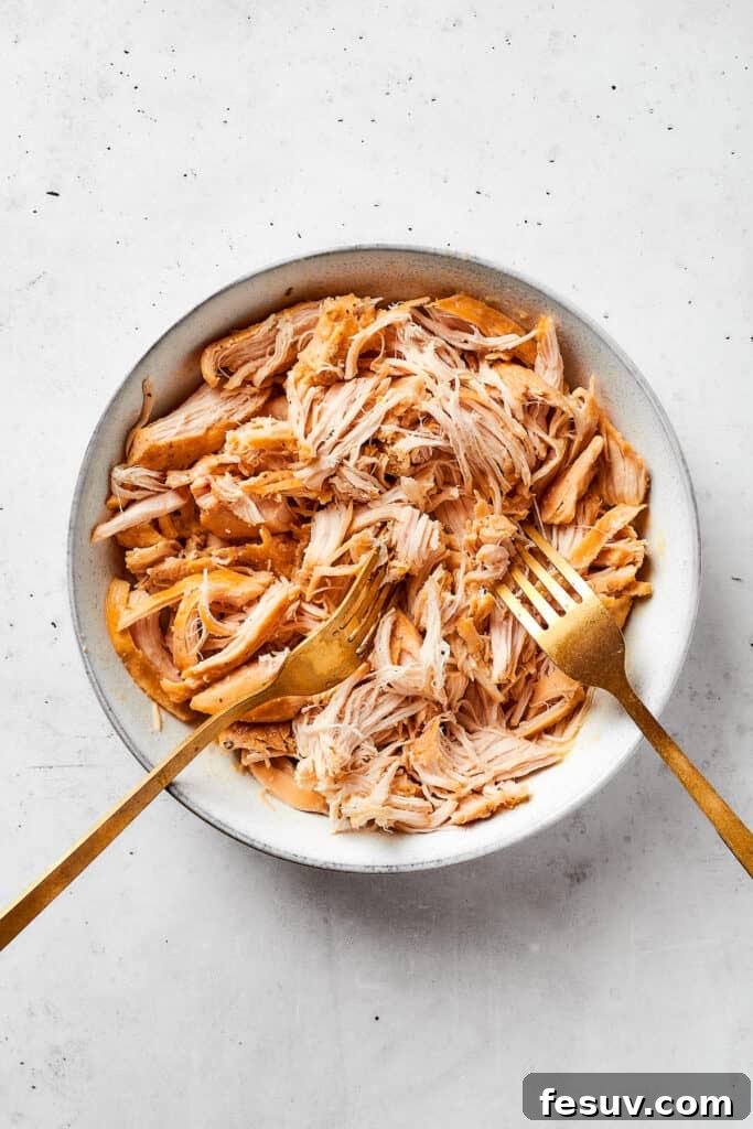 Two forks work to shred chicken in a white bowl.