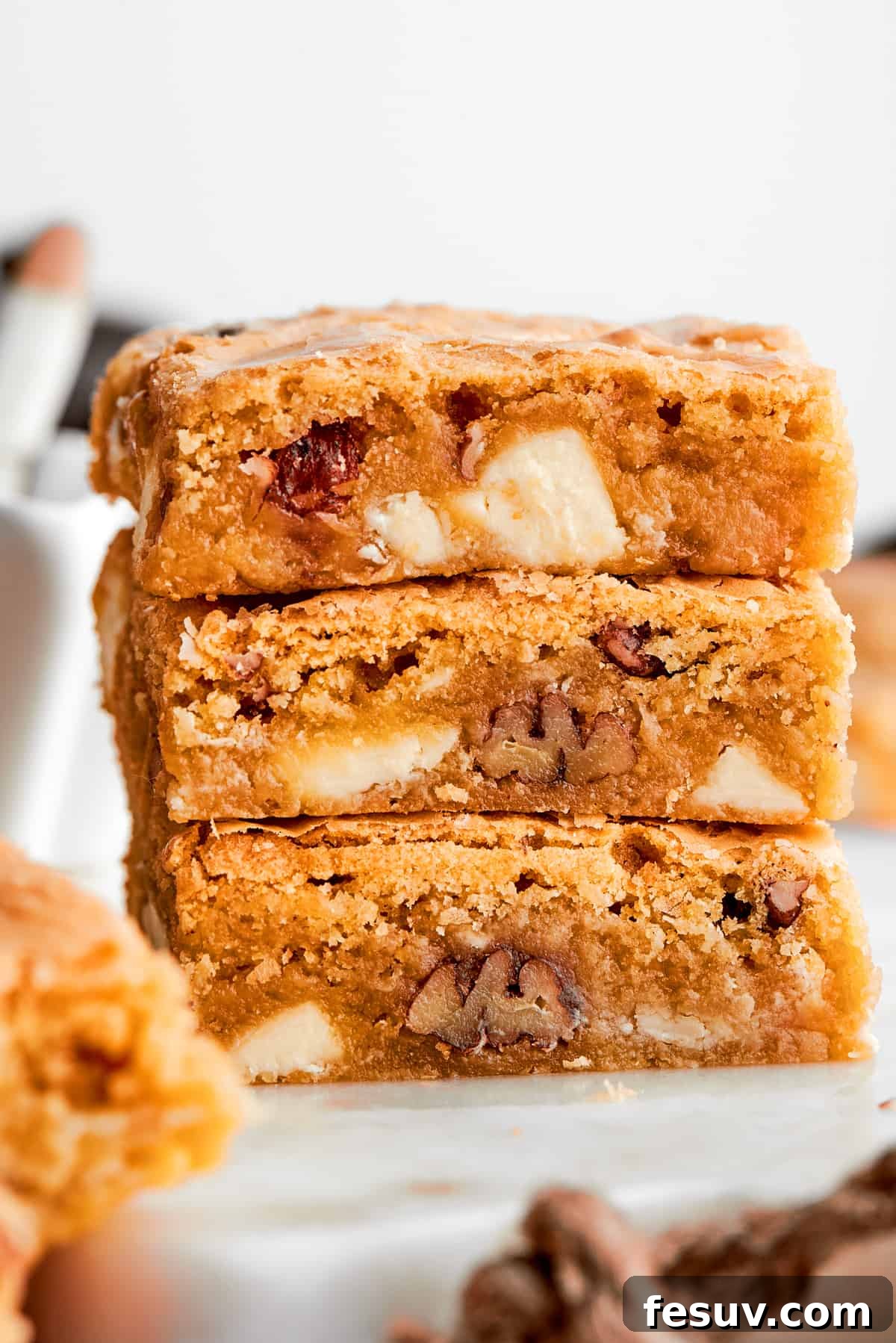 This sweet and irresistible Blonde Brownies recipe features blondies studded with white chocolate chips and crunchy pecans. A stack of three rich blonde brownies, revealing the perfect golden-brown crust, chewy vanilla interior, and generous scattering of white chocolate chips and crunchy pecans.