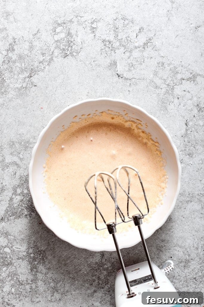 A hand mixer poised over the warmed egg mixture, ready to beat it until thick and cooled.
