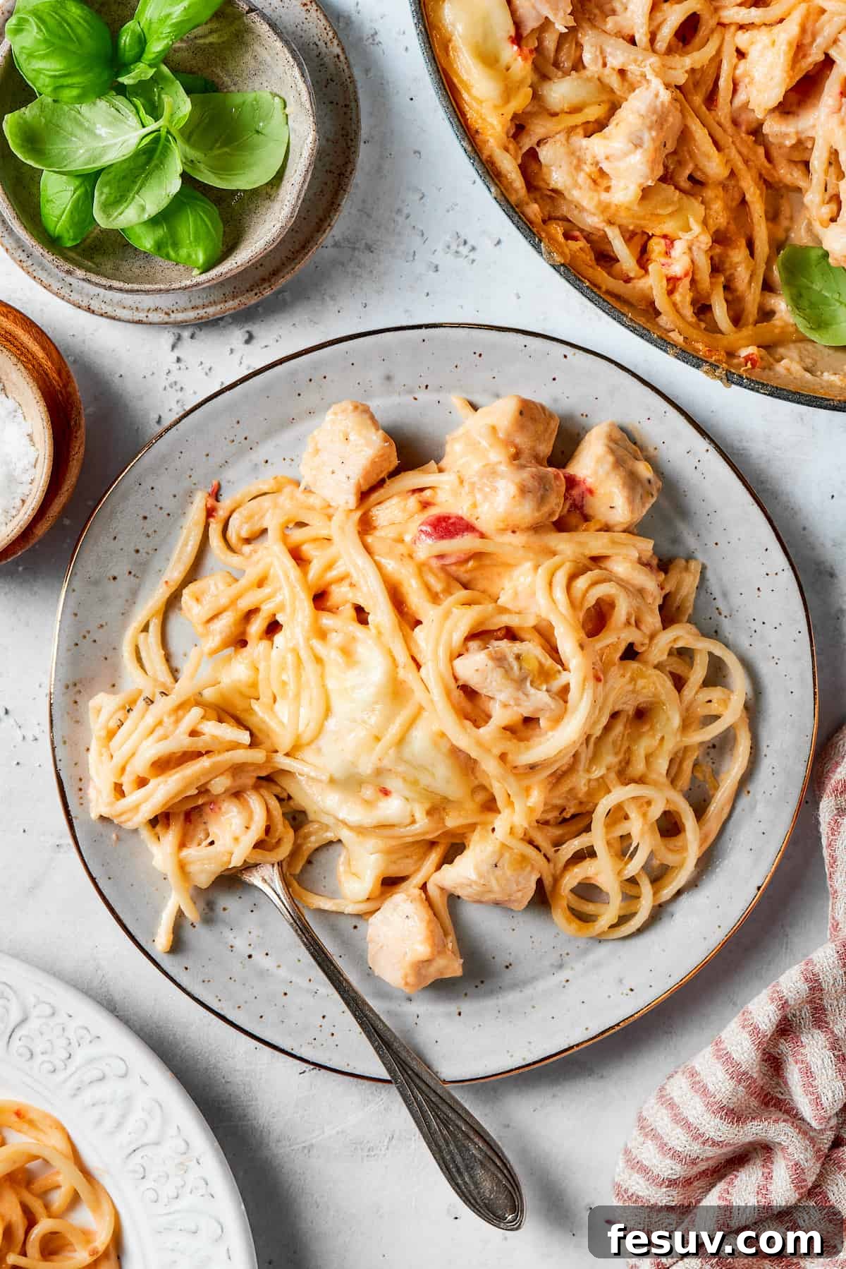 A plate of cheesy chicken spaghetti with a fork and a bowl of the spaghetti to the right, garnished with fresh basil.