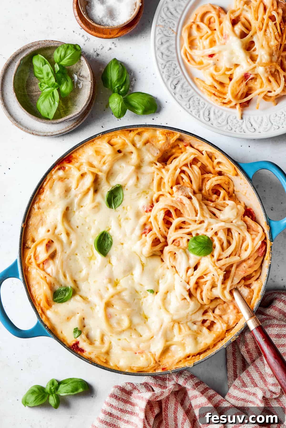 A pot of cheesy chicken spaghetti on a table with a bowl of fresh basil leaves next to it.