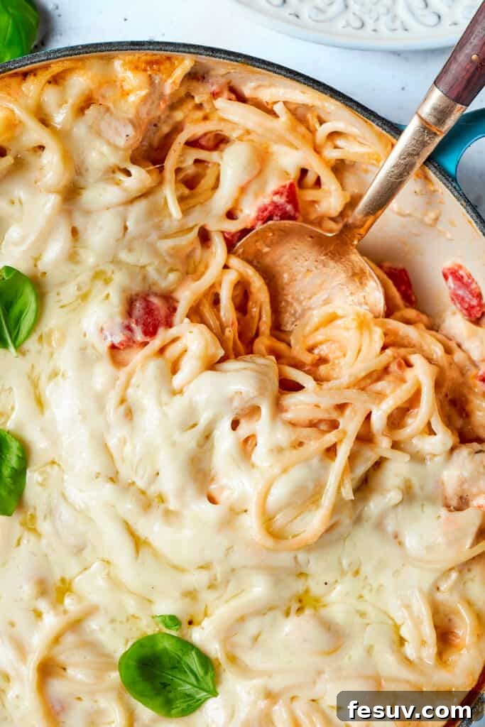 A spoon digs into a pot of cheesy chicken spaghetti, showing off the melted cheese and saucy pasta.