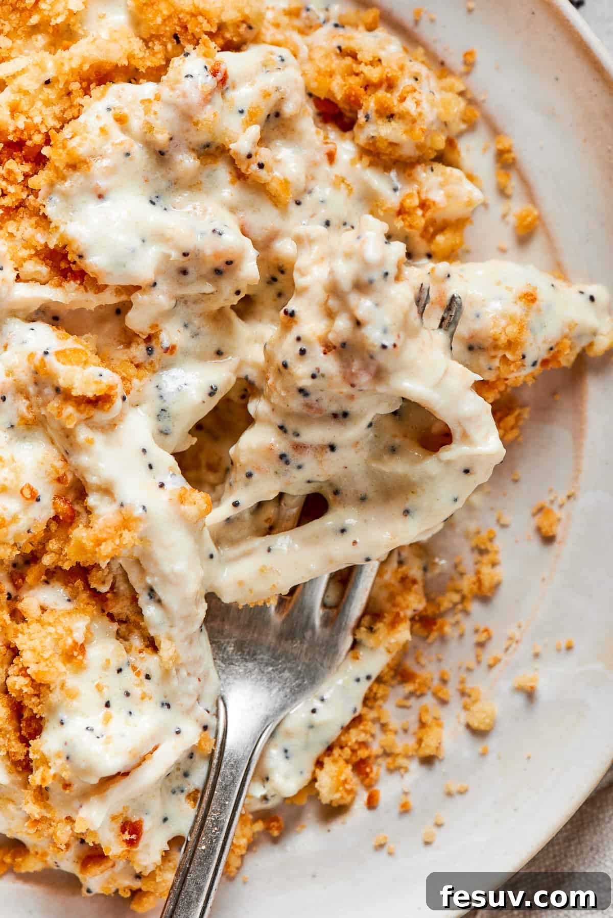 This creamy Poppy Seed Chicken casserole features a rich, poppyseed-flecked sauce that wraps cozily around tender shredded chicken. A fork lifts a succulent piece of poppy seed chicken, showcasing its creamy texture and the golden cracker topping, hovering above a plate already filled with a hearty serving of the delicious casserole.