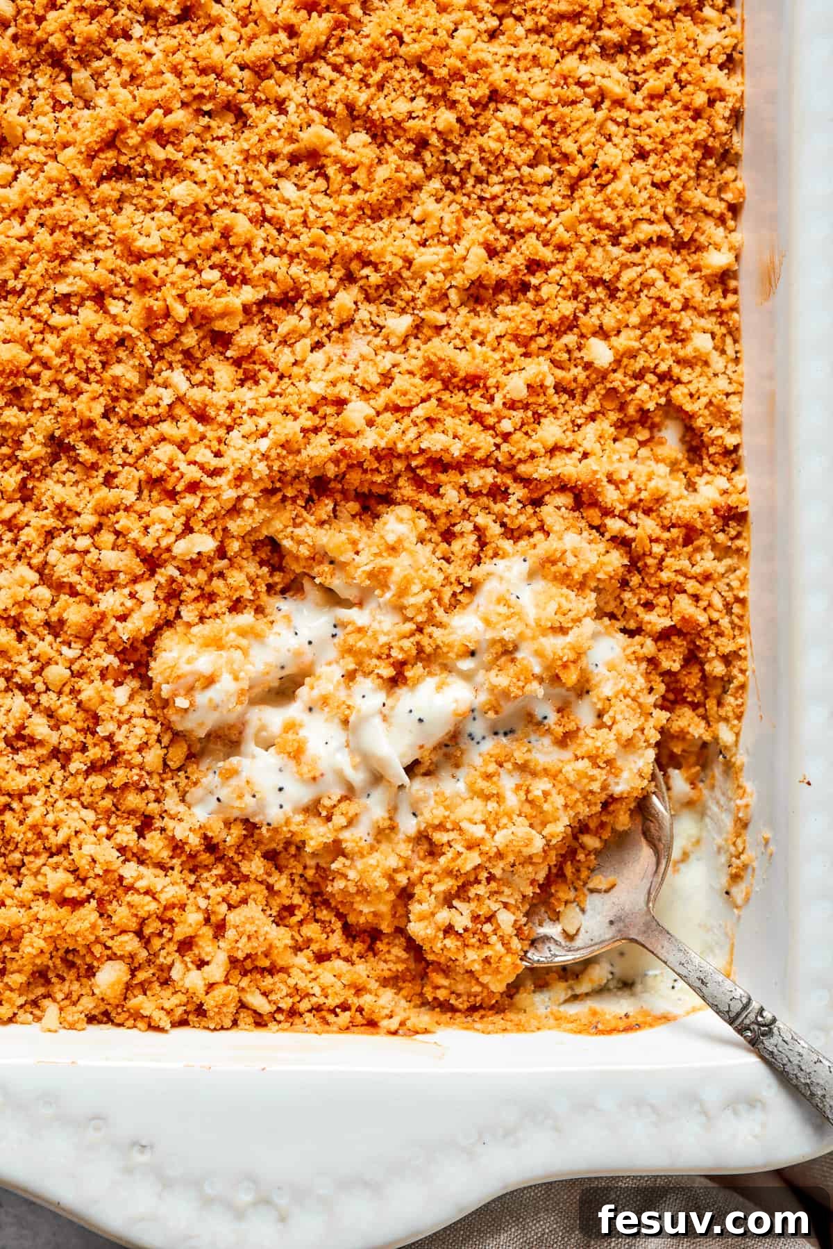 This creamy Poppy Seed Chicken casserole features a rich, poppyseed-flecked sauce that wraps cozily around tender shredded chicken. A large serving spoon delves into a bubbling dish of poppy seed chicken, revealing the creamy interior beneath a golden, crunchy topping of crushed Ritz crackers. The casserole is freshly baked and looks incredibly appetizing.