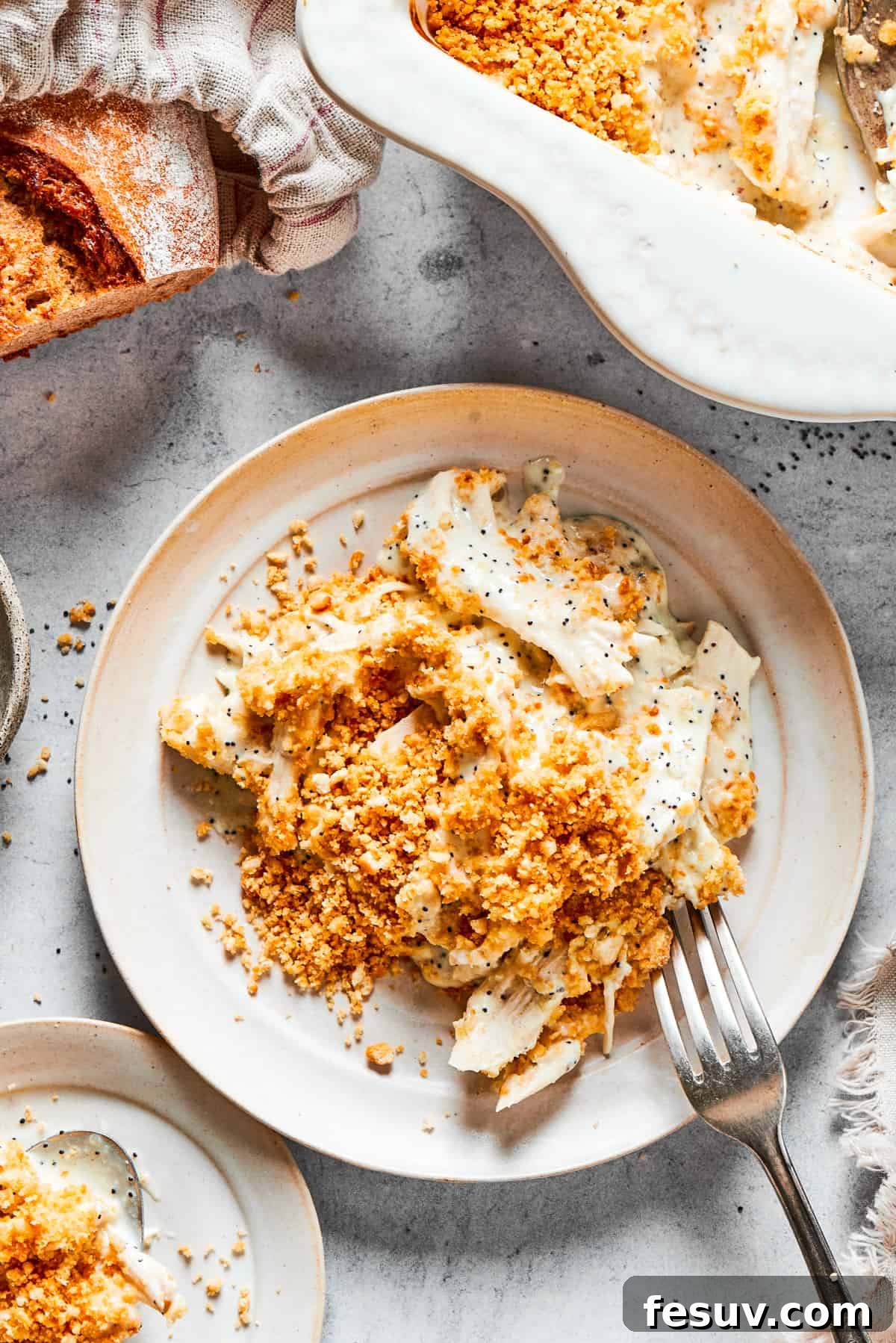 This creamy Poppy Seed Chicken casserole features a rich, poppyseed-flecked sauce that wraps cozily around tender shredded chicken. A beautifully presented casserole dish of poppy seed chicken, garnished and ready for serving on a rustic wooden table, accompanied by a loaf of fresh bread and a plate with a generous serving.