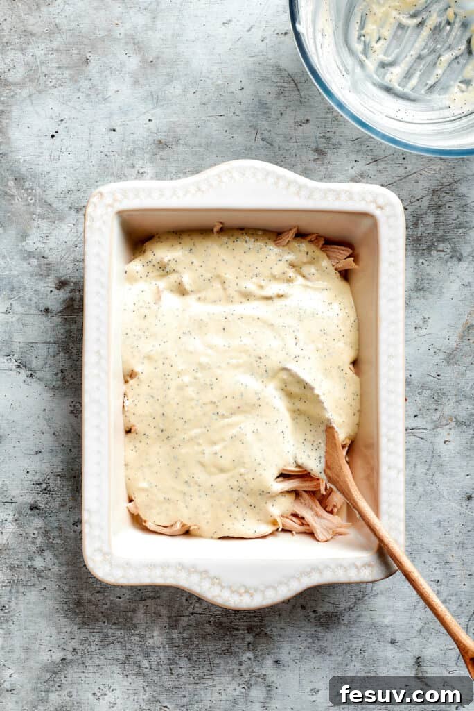 This creamy Poppy Seed Chicken casserole features a rich, poppyseed-flecked sauce that wraps cozily around tender shredded chicken. The creamy poppy seed sauce being smoothly spread over the layer of shredded chicken in a baking dish, ensuring even coating for a flavorful casserole.