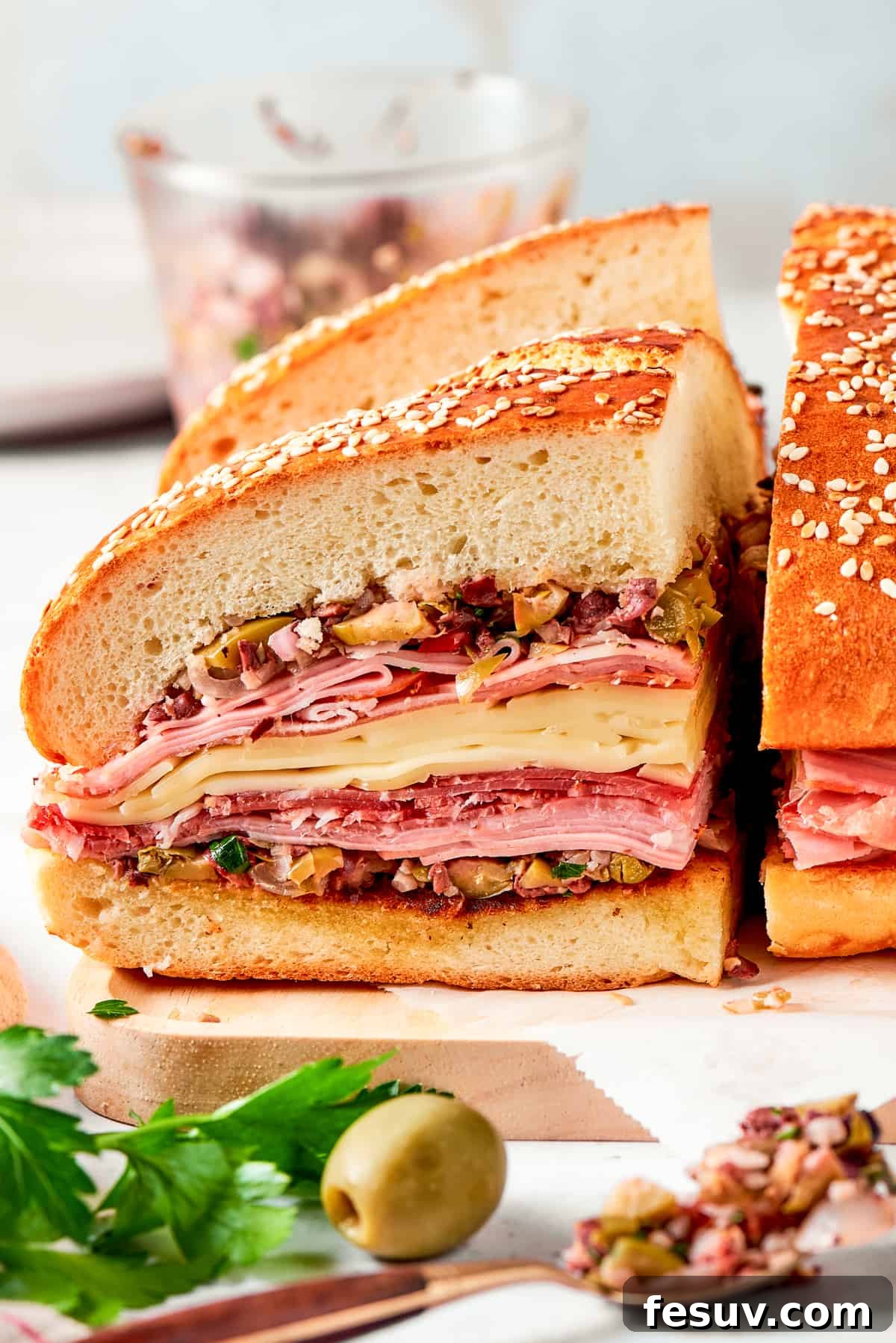 Two pieces of a tall, sliced muffaletta sandwich are presented on a wooden cutting board, showcasing the intricate layers of meat and cheese.
