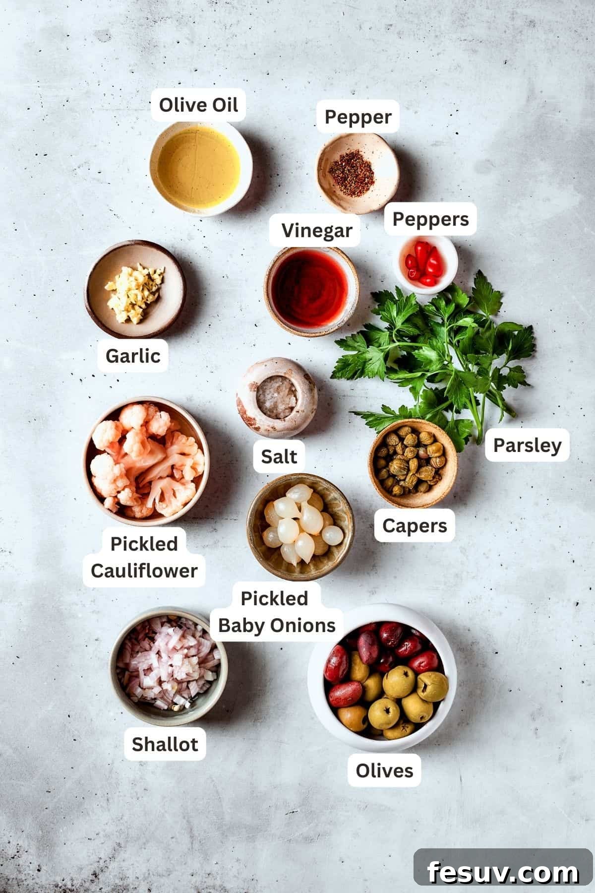 A vibrant array of ingredients for olive salad are displayed, including green olives, Kalamata olives, peppers, pickled cauliflower, capers, garlic, fresh parsley, and bottles of olive oil and red wine vinegar.