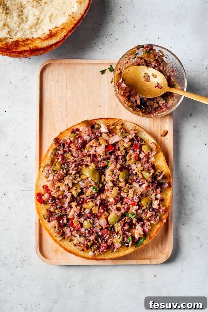 Freshly prepared olive salad being generously spread onto one half of the toasted muffaletta bread.