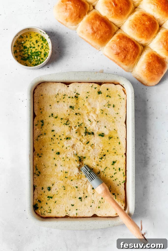 Applying the flavorful butter mixture to the rolls. A vibrant melted butter mixture is being expertly brushed onto the bottom halves of sweet Hawaiian rolls, which are neatly arranged in a baking dish, preparing them to absorb the rich, savory flavors.