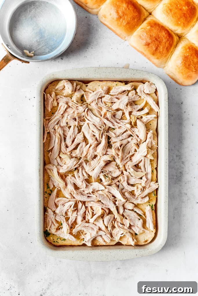 Layering the shredded chicken evenly onto the rolls. Tender shredded chicken is generously and evenly spread over the buttered Hawaiian rolls in the baking dish, forming the flavorful and hearty base of the slider filling.