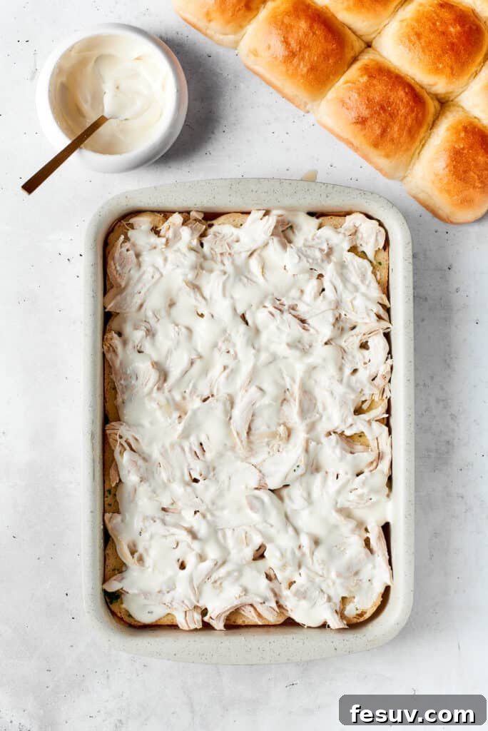 Adding the creamy ranch and a layer of cheese. A creamy, luscious mixture of ranch dressing is being skillfully drizzled over the shredded chicken and bacon, followed by a generous layer of melted cheese, meticulously preparing the sliders for the subsequent layers.