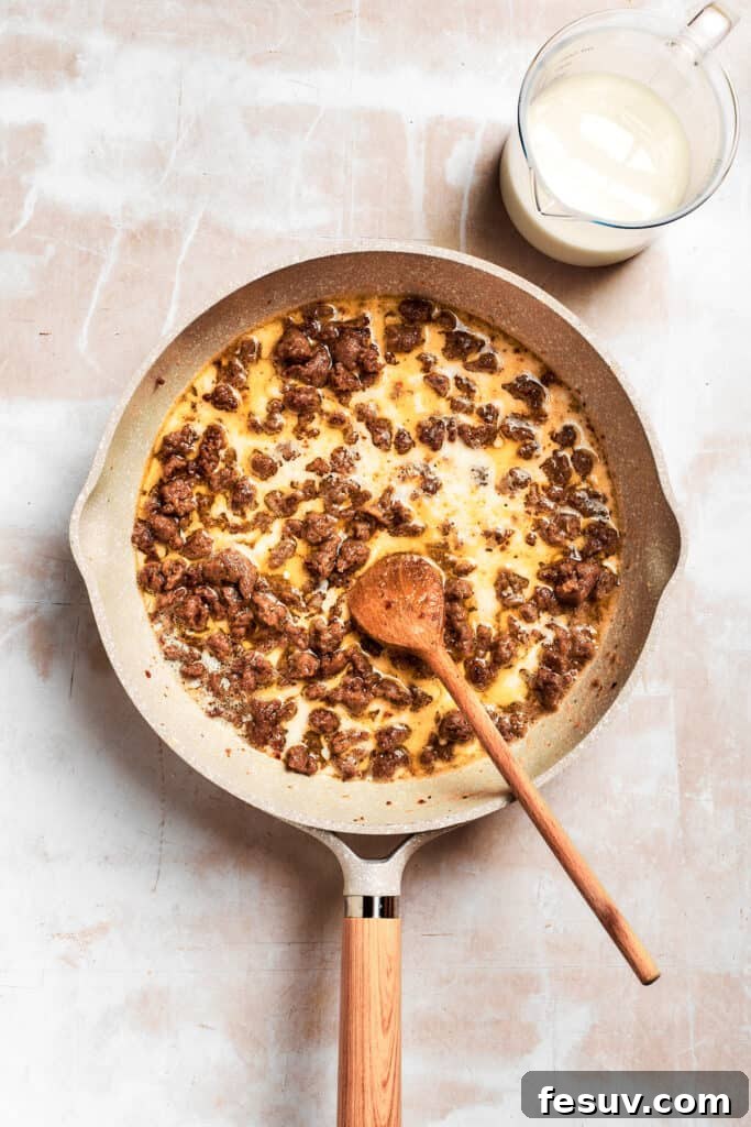 The sausage is stirred in the pan. The sausage and roux mixture being stirred vigorously in the skillet as milk is gradually added, transforming it into a smooth, thickening gravy.