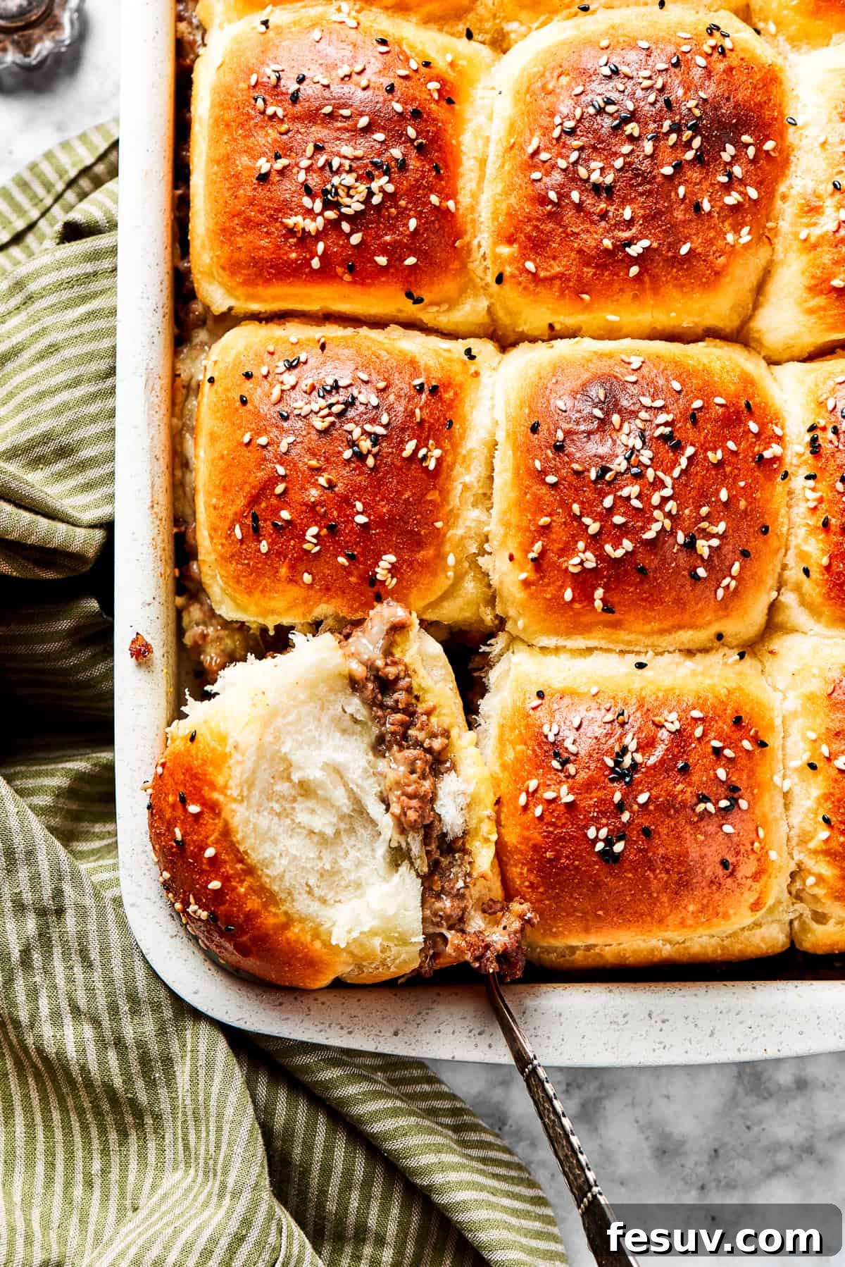 A dish of perfectly baked cheeseburger sliders, with one slider lifted to show its cheesy, meaty filling.