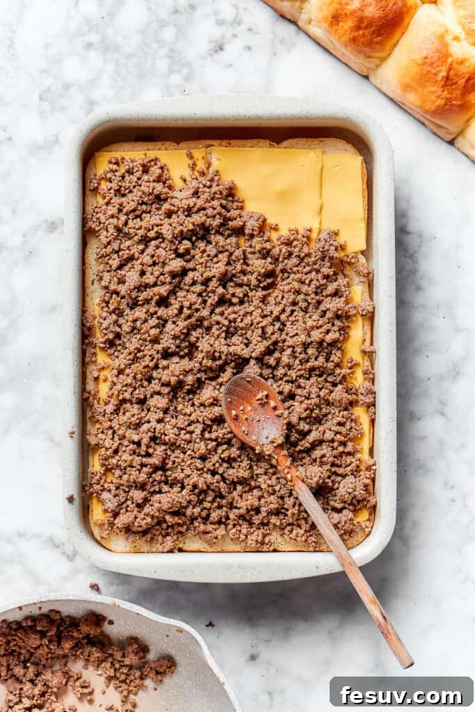 The seasoned ground beef mixture being spread evenly over the cheese layer on the dinner rolls.