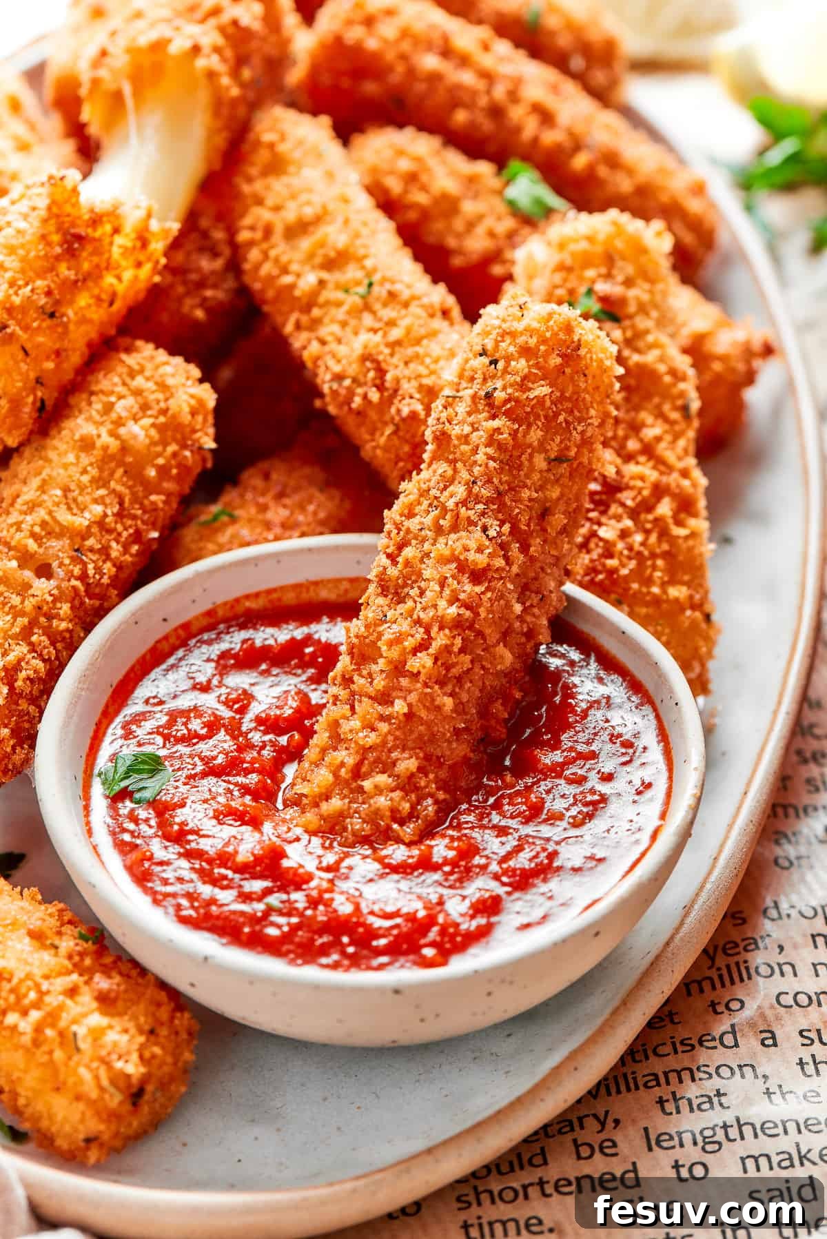 A golden-brown mozzarella stick is being dipped into a small, elegant bowl of vibrant red marinara sauce, showcasing its perfect crispness.