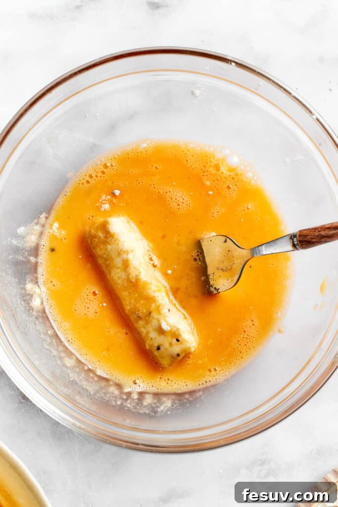 A mozzarella stick, already coated in flour and egg once, is being dipped again into the beaten egg for a second coating layer.