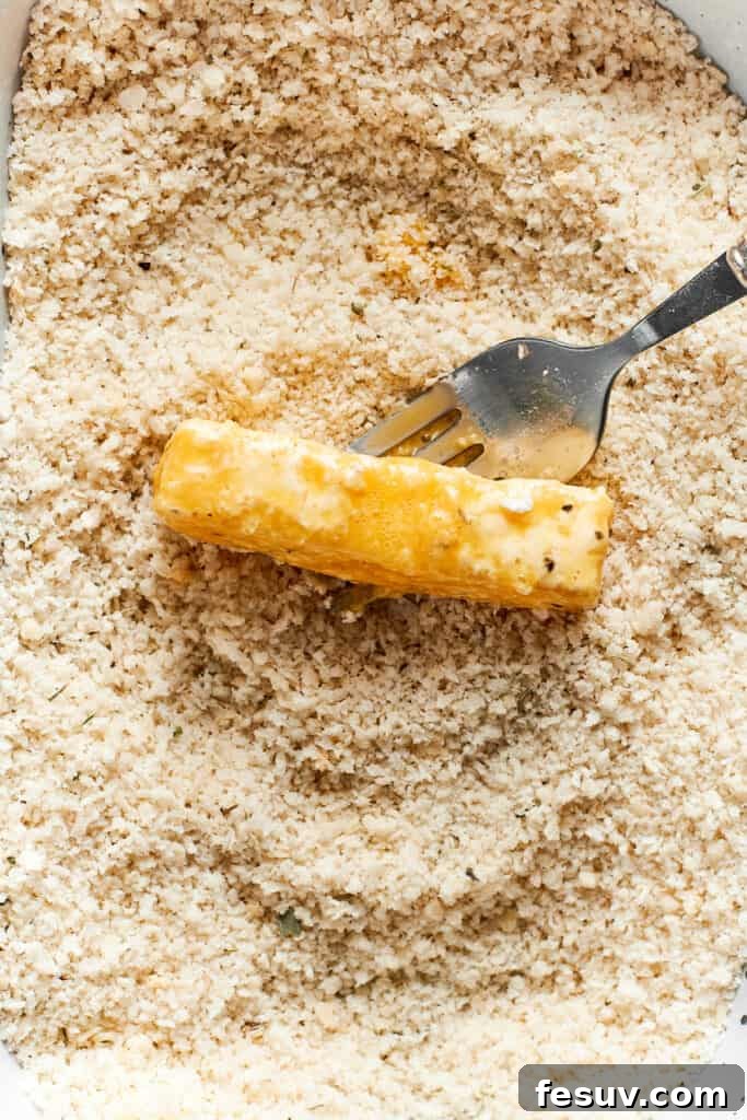 A fork is used to gently toss a double-coated mozzarella stick in a dish of seasoned panko breadcrumbs, ensuring full coverage.