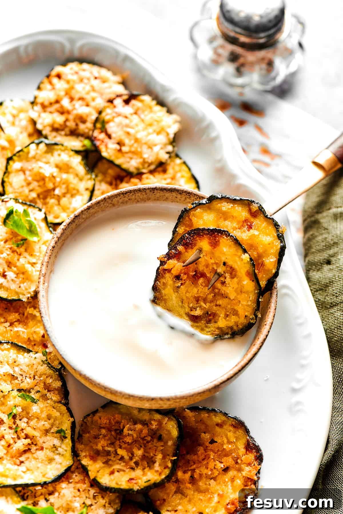Two golden, crispy zucchini chips are dipped into a small bowl of creamy yogurt, which sits on a larger platter with more chips.