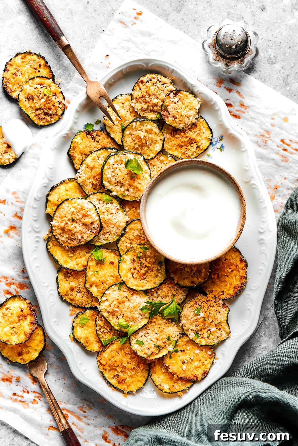 A white oval platter filled with golden, crispy baked zucchini chips, accompanied by a small bowl of creamy dip and fresh herb garnish.