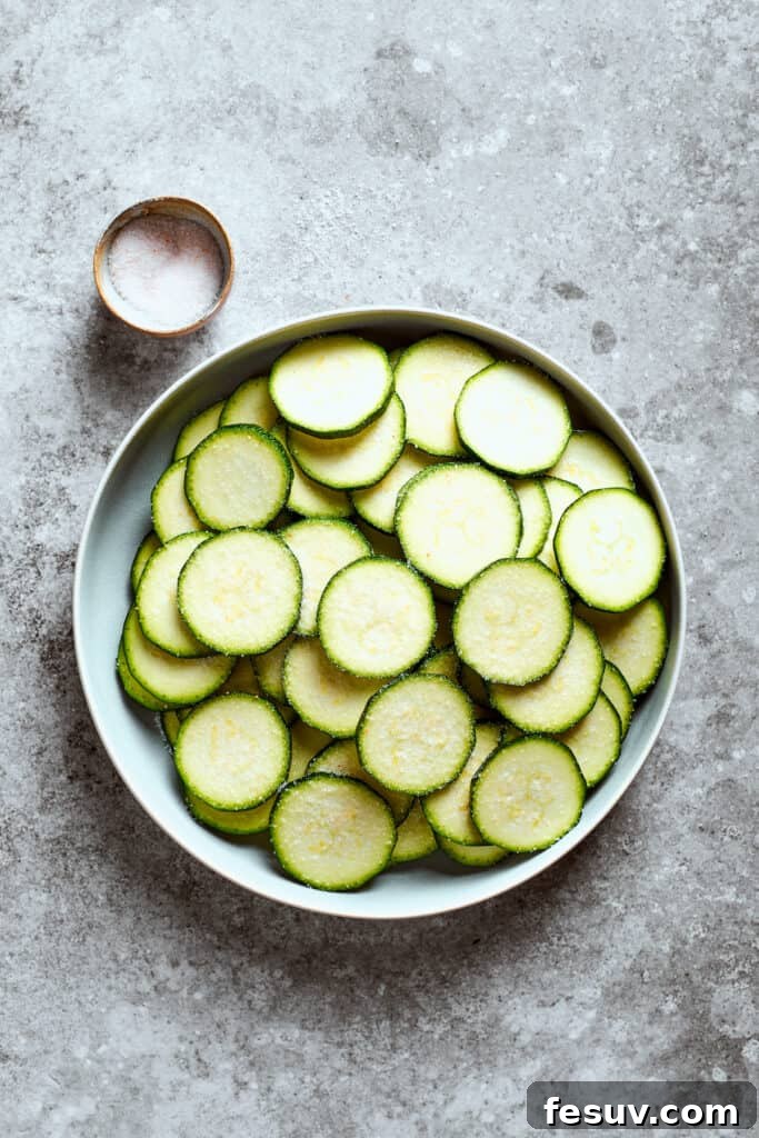 A bowl of uniformly sliced zucchini rounds sits beside a small dish of sea salt, ready for the draining process.
