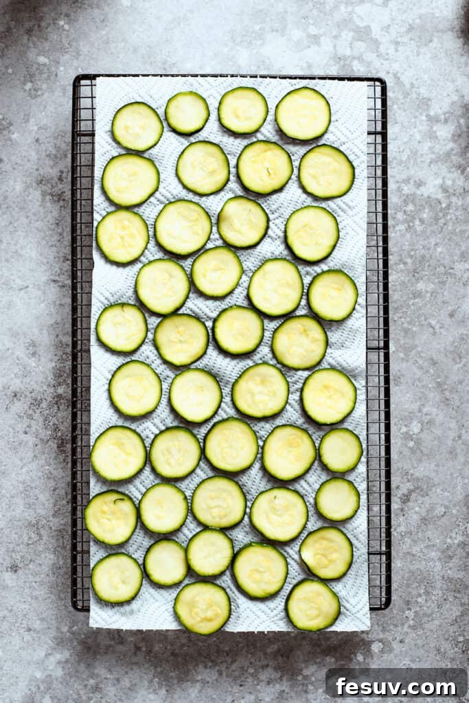 Numerous thin zucchini slices are neatly arranged in a single layer on a large baking sheet, ready for the next step.