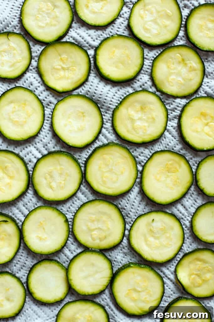 Thin zucchini slices are spread out on a baking sheet, glistening slightly after being prepared.