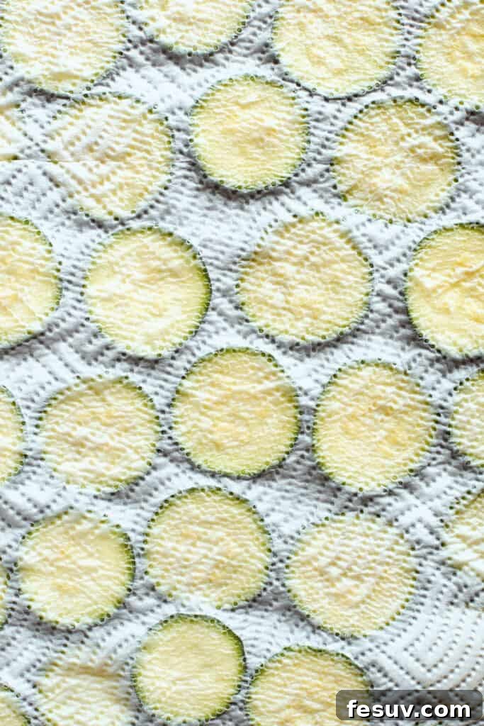 A baking sheet of zucchini slices is covered with a paper towel, absorbing excess moisture for crisper chips.