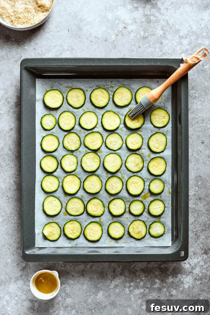 A pastry brush applies olive oil evenly over thinly sliced zucchini rounds arranged on a baking sheet.
