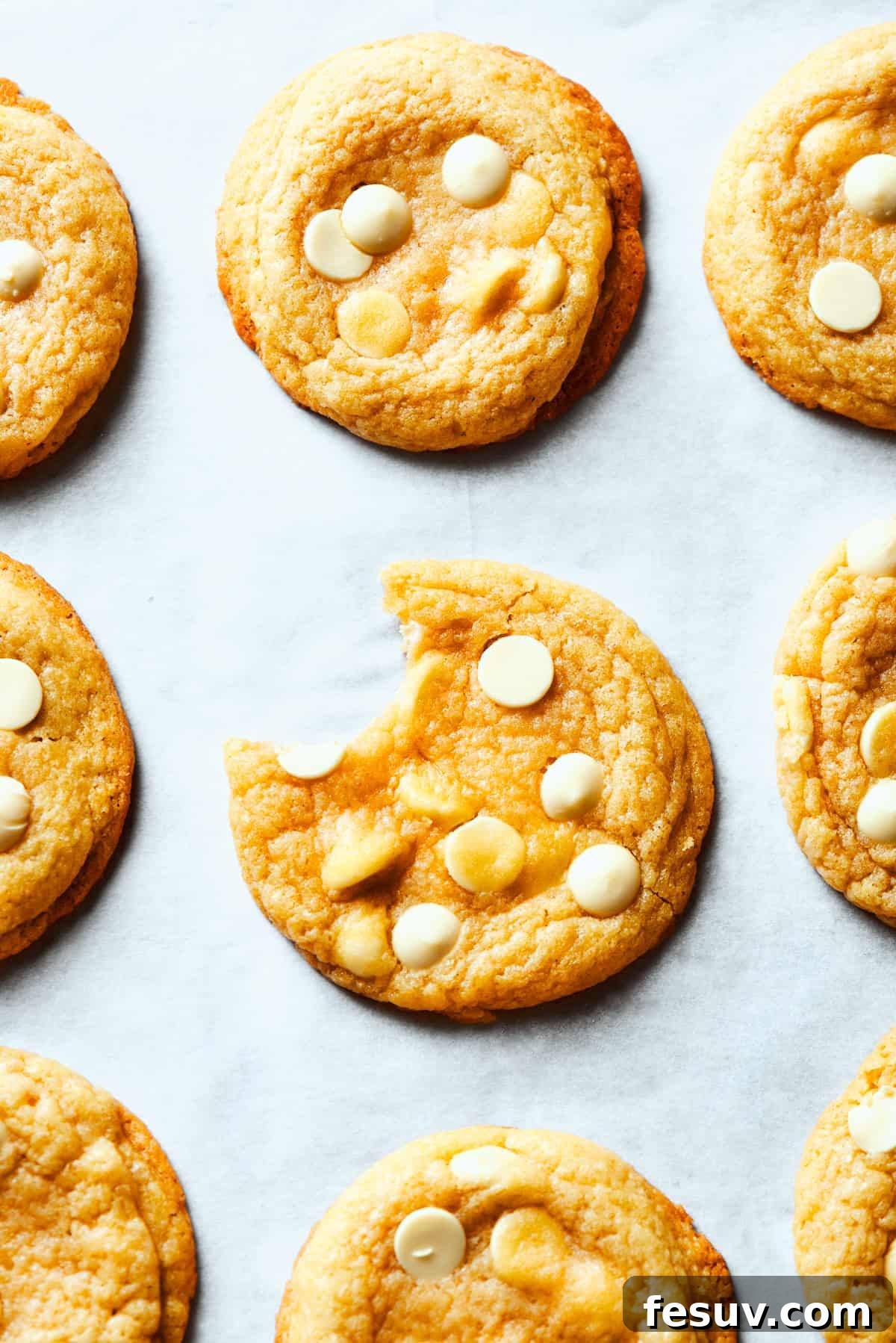 Chewy, buttery White Chocolate Chip Cookies are packed with creamy, sweet white chocolate chips instead of semisweet for an unexpected twist. A perfectly baked white chocolate chip cookie sits in the center of a rustic white plate, with a bite missing, revealing its soft, chewy interior filled with melted white chocolate chips. Other cookies are artfully arranged around it.
