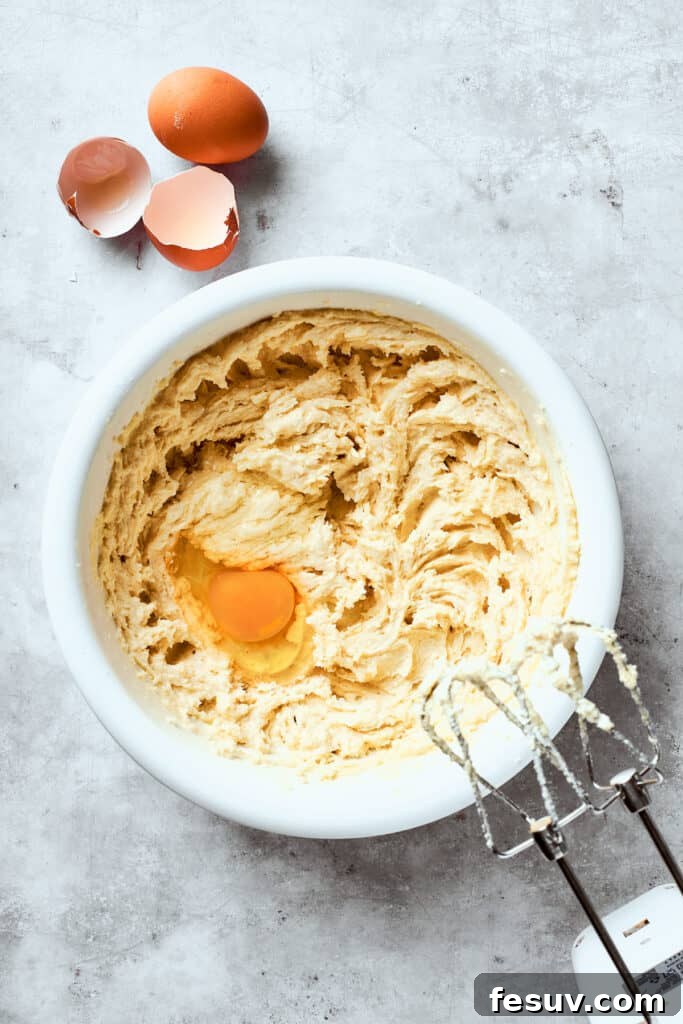 Chewy, buttery White Chocolate Chip Cookies are packed with creamy, sweet white chocolate chips instead of semisweet for an unexpected twist. A fresh large egg is being cracked and beaten into the creamy butter and sugar mixture in a large mixing bowl, as part of the process for making white chocolate chip cookie dough.