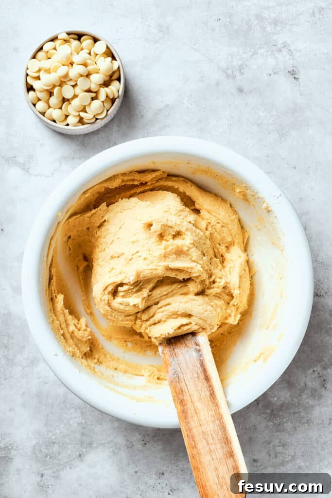 Chewy, buttery White Chocolate Chip Cookies are packed with creamy, sweet white chocolate chips instead of semisweet for an unexpected twist. The white chocolate chip cookie dough, a rich, buttery mixture, is being gently stirred in a large mixing bowl with a spatula, ensuring all ingredients are just combined and evenly incorporated.