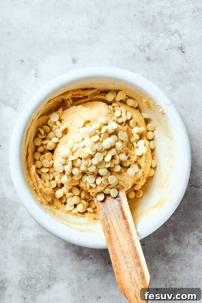 Chewy, buttery White Chocolate Chip Cookies are packed with creamy, sweet white chocolate chips instead of semisweet for an unexpected twist. Creamy white chocolate chips are being gently folded into the prepared cookie dough in a large mixing bowl, ensuring even distribution throughout the rich, buttery mixture.
