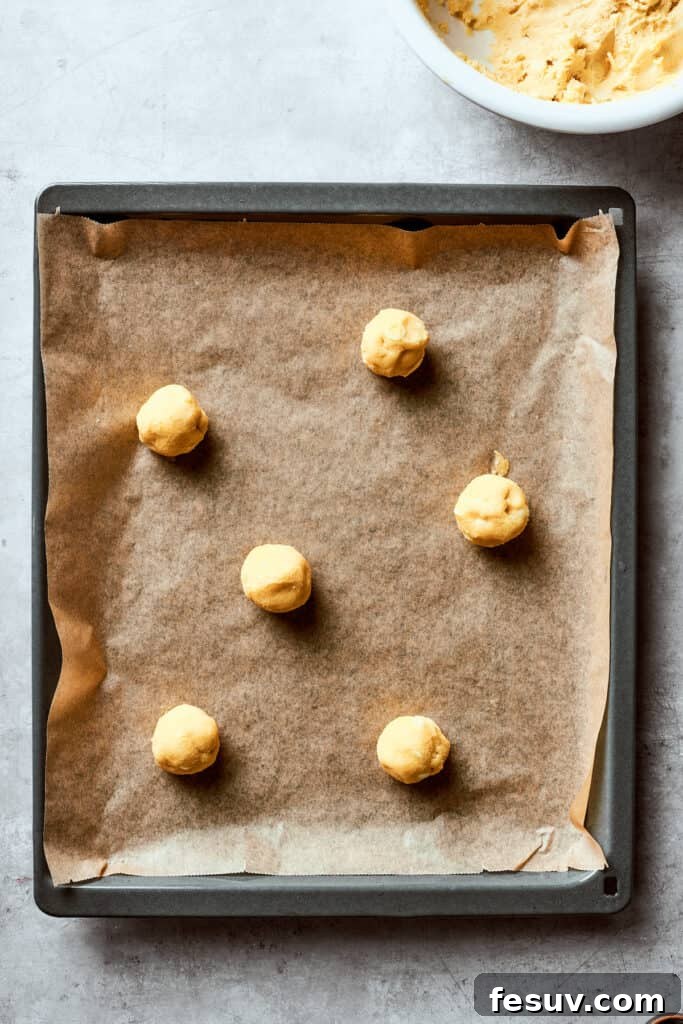Chewy, buttery White Chocolate Chip Cookies are packed with creamy, sweet white chocolate chips instead of semisweet for an unexpected twist. Uniform balls of white chocolate chip cookie dough are neatly arranged on a parchment-lined baking sheet, leaving ample space between each for even spreading during baking, ready to be placed in the oven.