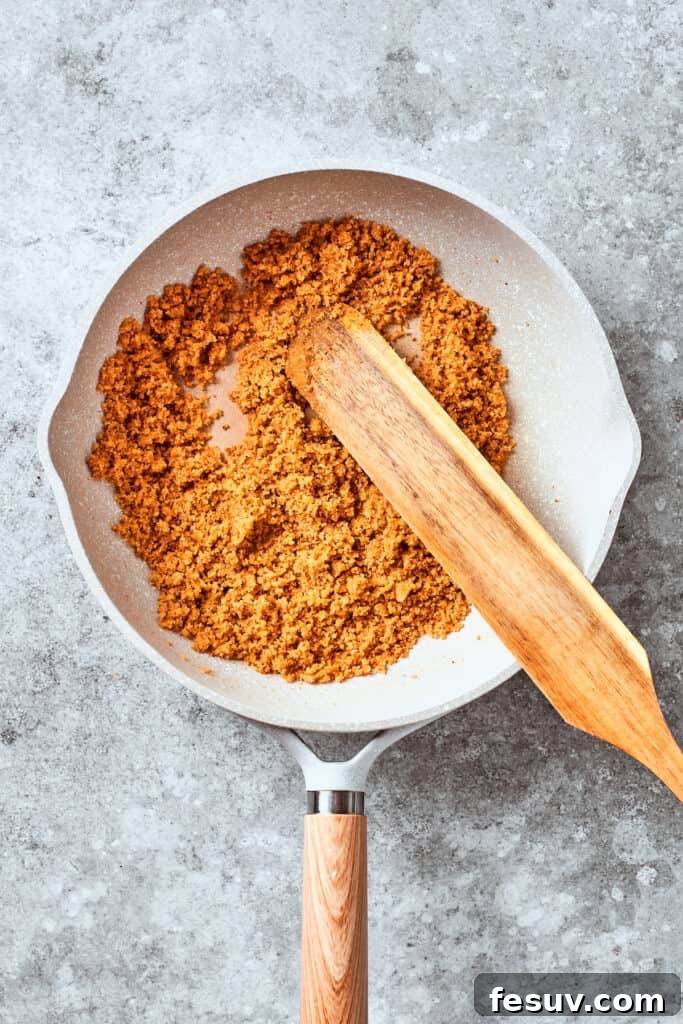 A wooden spoon is stirring Panko breadcrumbs in a hot pan, toasting them to a perfect golden brown for the mac and cheese topping.