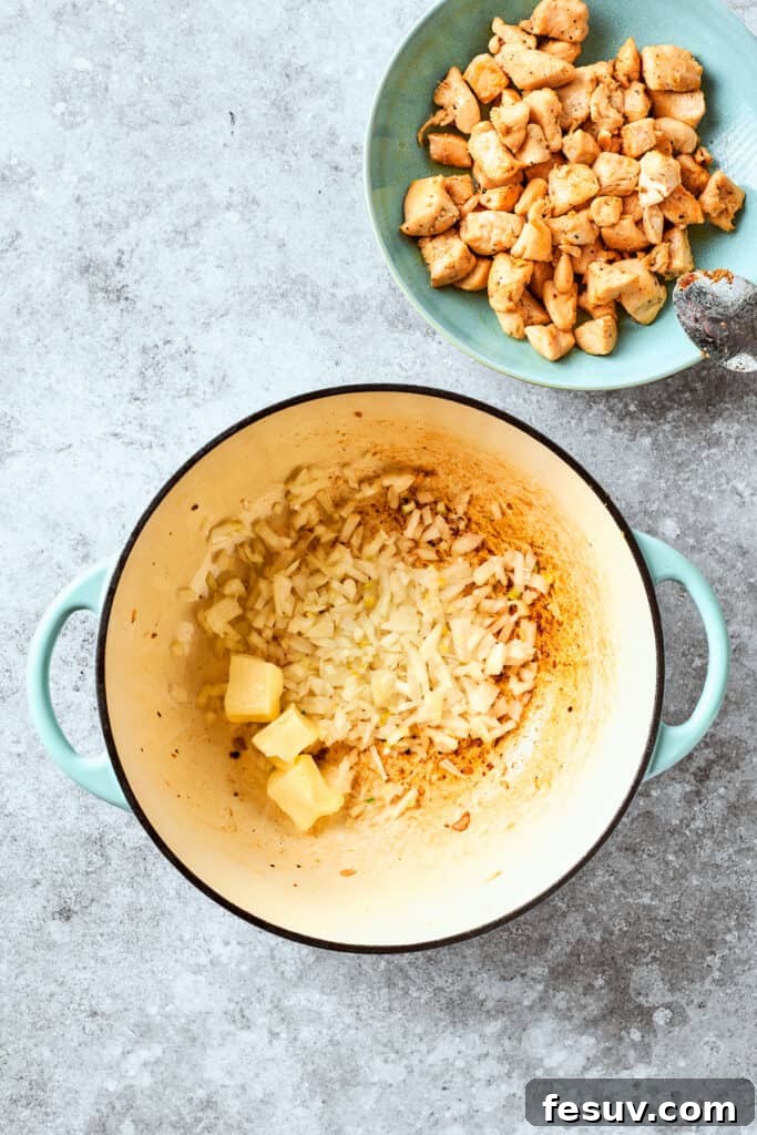 Finely chopped onion and butter are sautéing in a skillet next to a bowl of cooked chicken cubes, ready to build the sauce base.